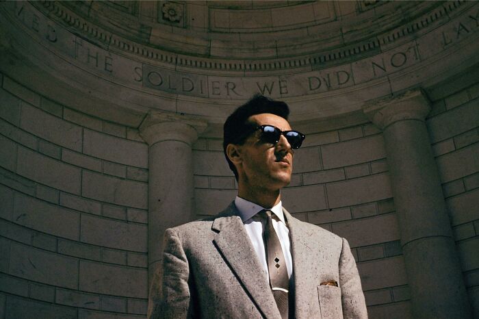 Man in a gray suit and sunglasses embodying 60s fashion looks, standing in front of an ornate stone structure.
