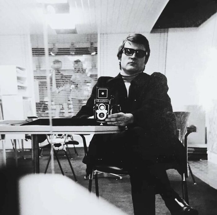 Man in 60s fashion with sunglasses and suit, holding a vintage camera, reflected in a modern setting.
