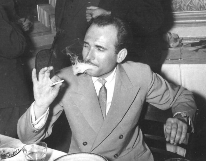 Man in a classic double-breasted suit from 60s fashion looks, smoking at a dinner table, showcasing vintage style.