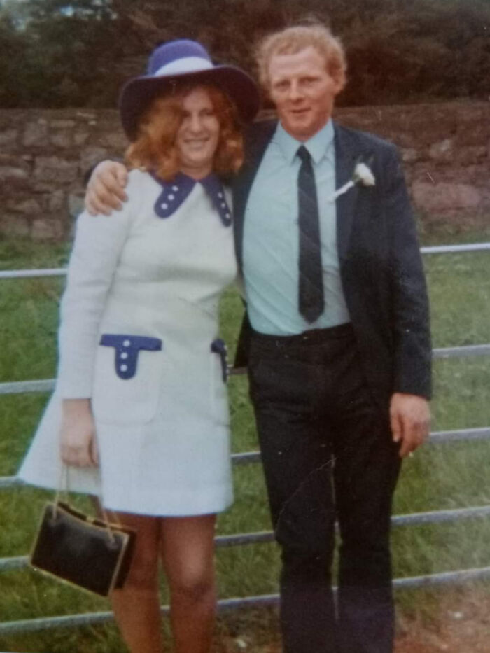 Vintage couple in 60s fashion, woman in white dress and blue hat, man in suit with tie, smiling outdoors.