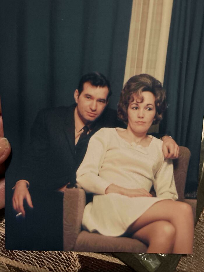 A couple in 60s fashion, man in a suit holding a cigarette, woman in a cream dress seated, capturing vintage style elegance.