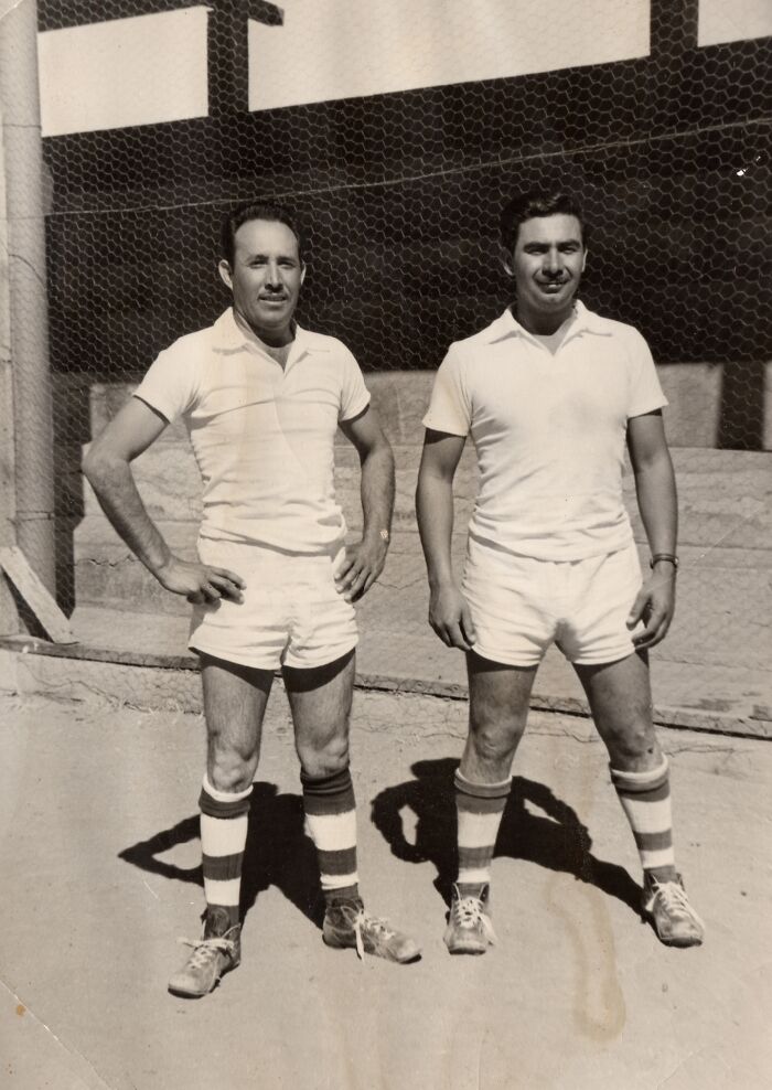 Two men in 60s fashion, wearing sporty white outfits and striped socks, standing outdoors.
