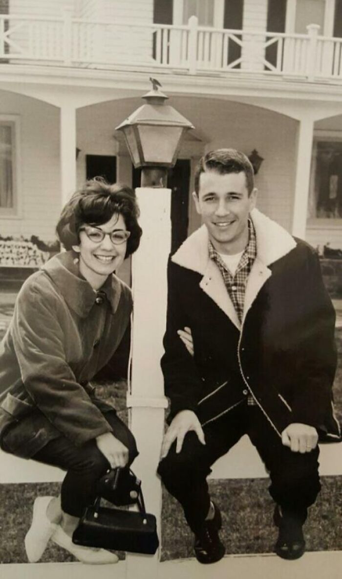 A smiling couple in classic 60s fashion, with the woman in glasses and coat, and the man in a lined jacket, leaning by a lamp post.