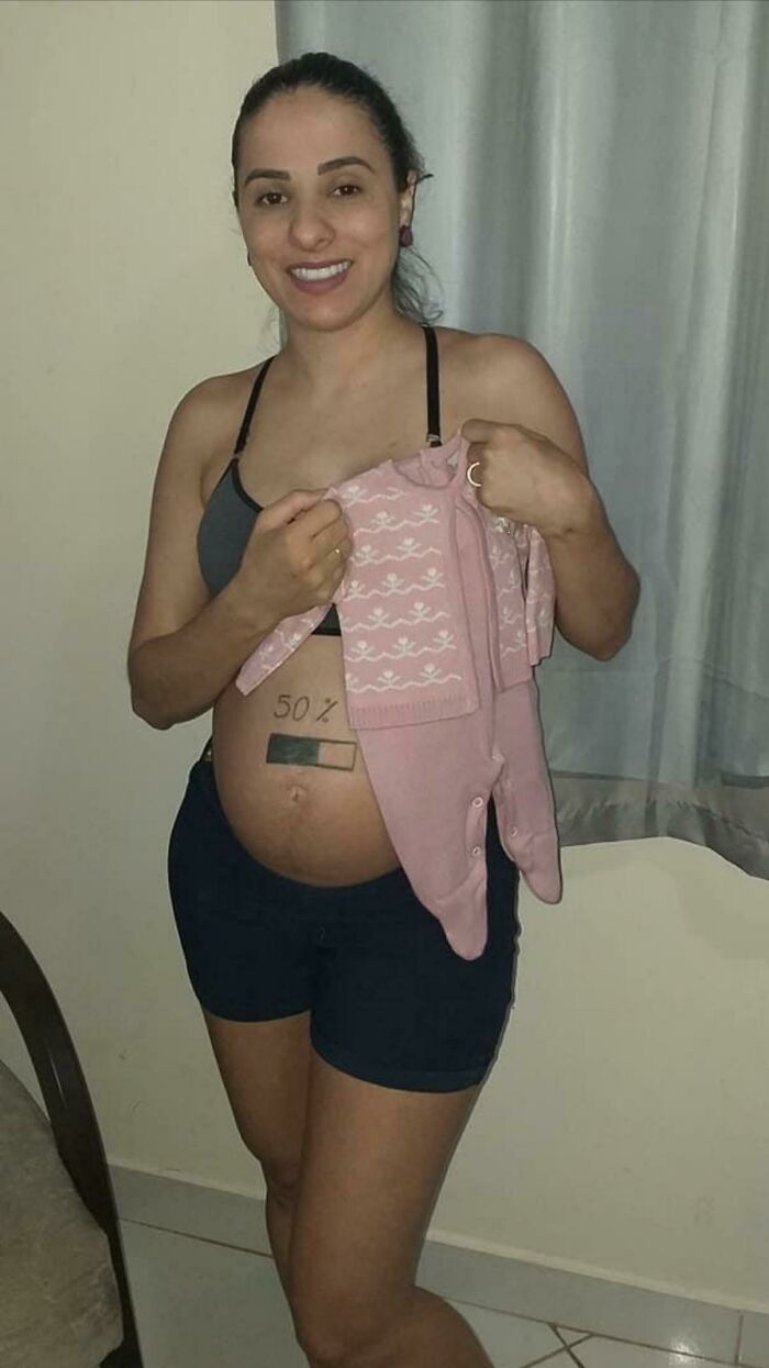 Pregnant woman humorously displays "50%" drawn on her belly while holding a pink baby outfit.