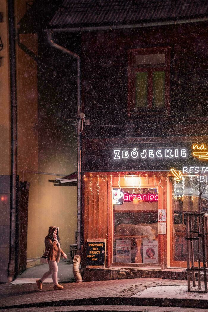 A person with a hood walks past a brightly lit restaurant at night, capturing a creative street photography moment.