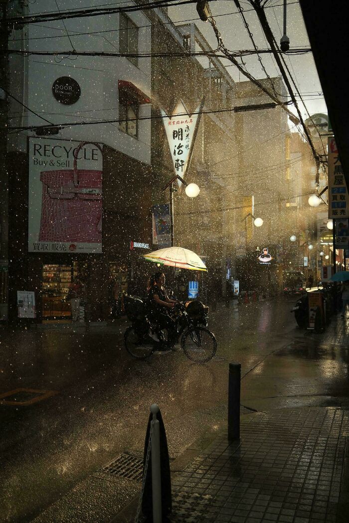 Cyclist with umbrella in rainy city street, showcasing creative street photography with dramatic lighting.