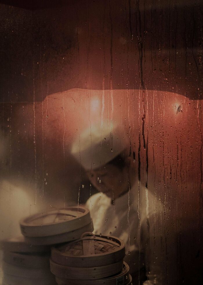Chef behind a steamy window with dim sum baskets, capturing creative street photography ambiance.
