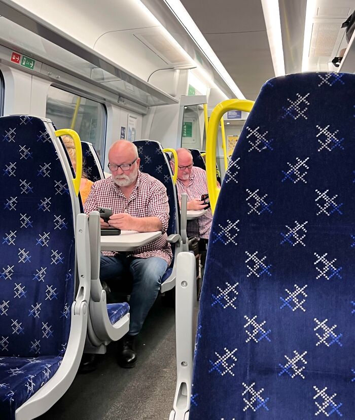 Man sitting on a train wearing glasses, holding a phone, with another man in similar attire seated behind him. Fascinating pics.