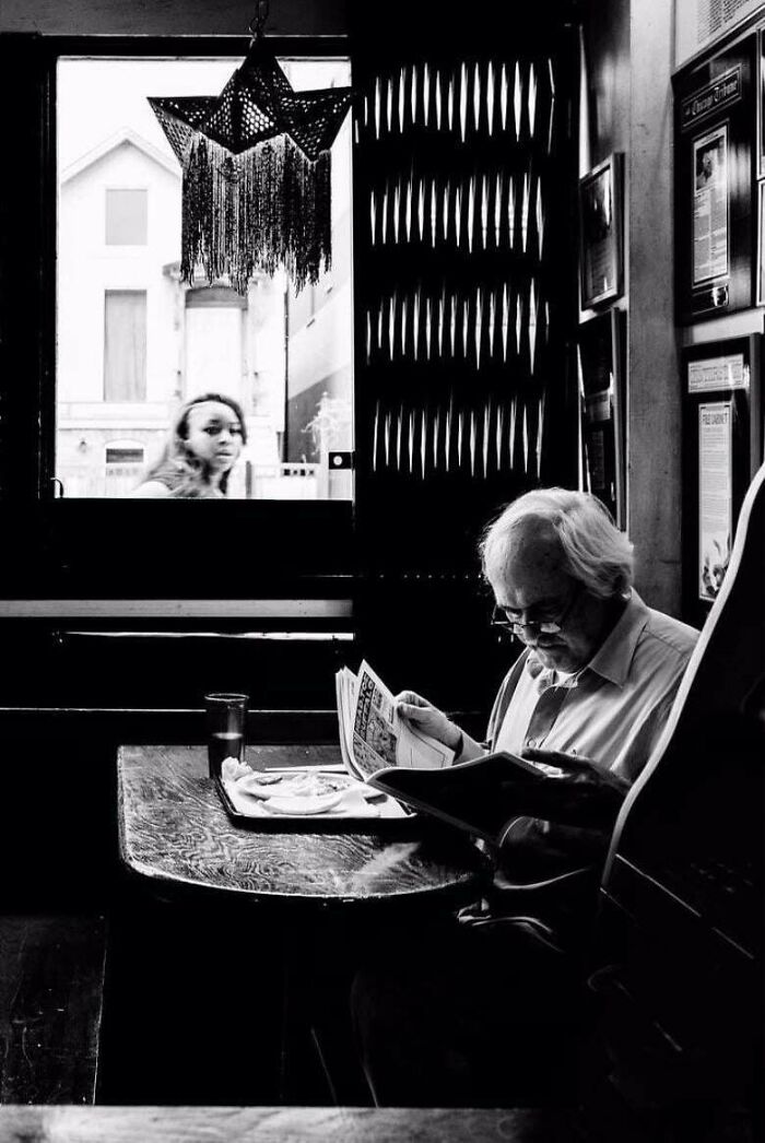 A man reads a newspaper inside a café, while a woman walks by the window, capturing a moment in creative street photography.