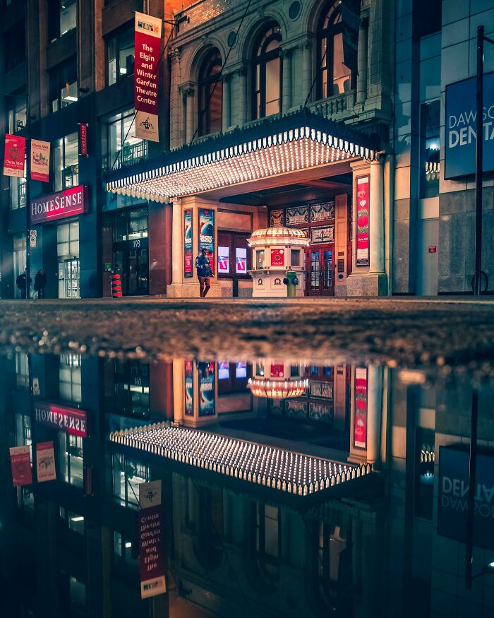 Creative street photography capturing a city theatre at night, with vibrant lights and reflections in a puddle.