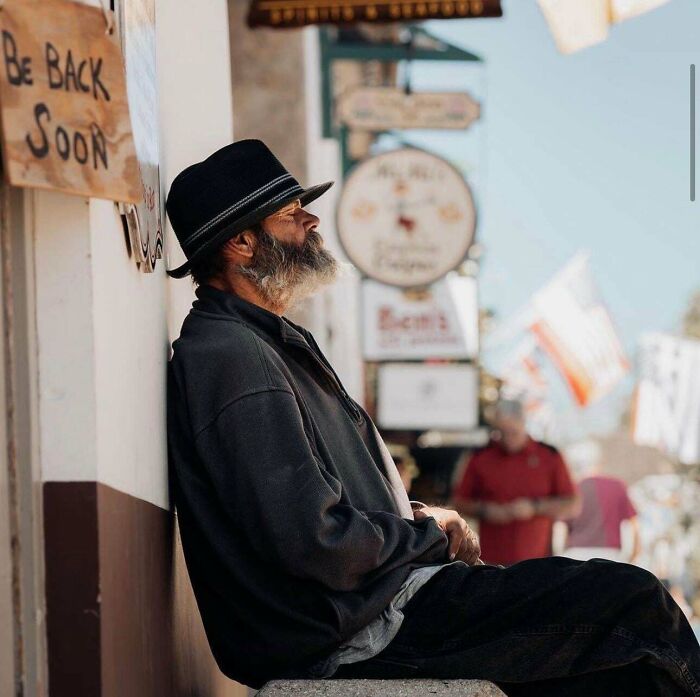 Man in a hat leans against a wall beside a "Be Back Soon" sign, showcasing creative street photography.
