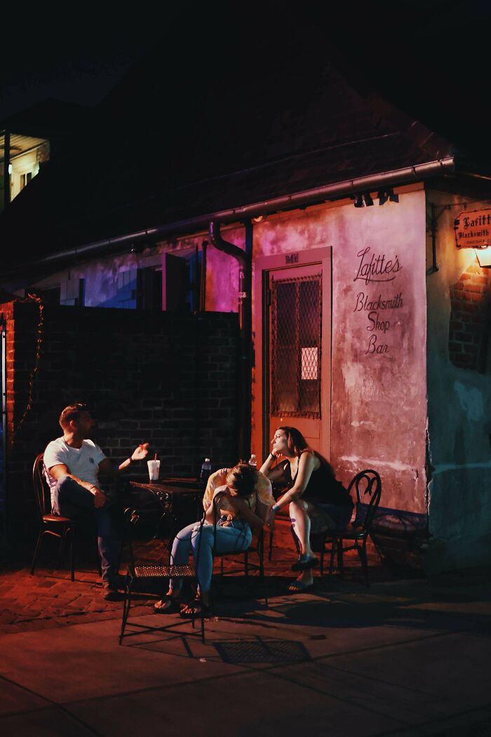 Three people sitting outside a bar at night, engaging in candid conversation. Creative street photography captures the moment.