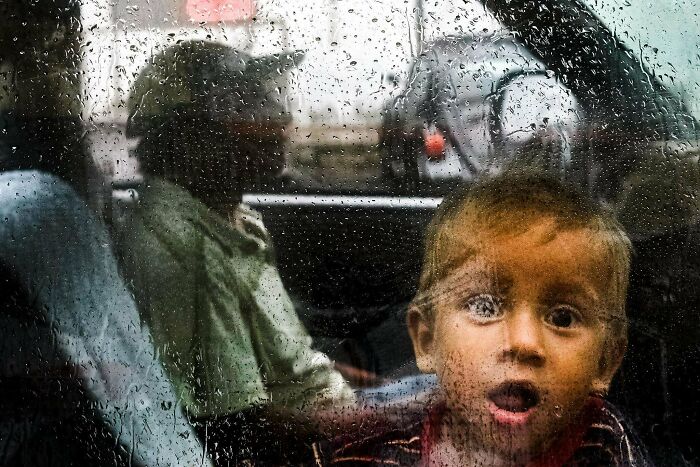 Child looking through a rain-covered window with a person sitting in the background, illustrating creative street photography.