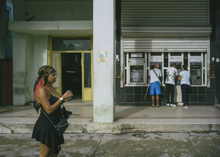 Woman in a black dress with a camera, three people at ATMs; scene captures creative street photography.