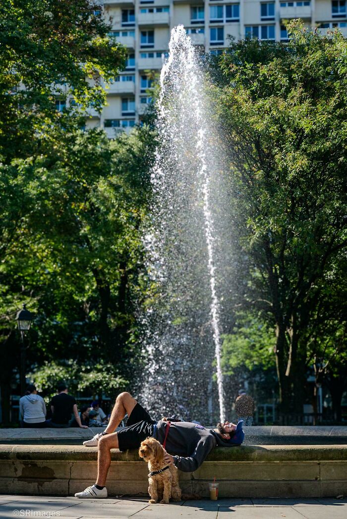 Man relaxing by a fountain with his dog, showcasing creative street photography in an urban park setting.