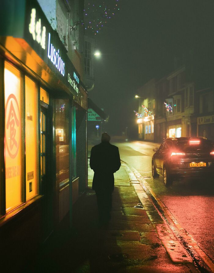 A silhouetted figure walks on a rain-soaked street at night, with illuminated shop signs and car lights—creative street photography.
