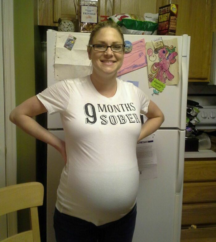 Smiling pregnant woman wearing a "9 Months Sober" T-shirt, standing in a kitchen.