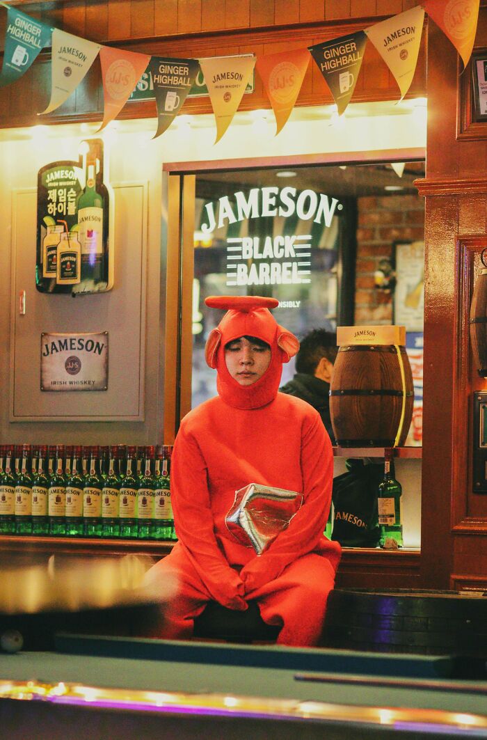 Person in red costume sitting in a bar, surrounded by whiskey bottles, reflecting creative street photography.