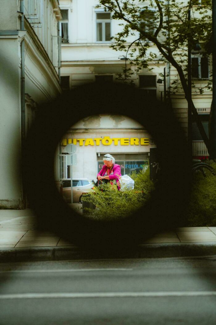 Woman in pink coat stands on street, viewed through a circular frame, showcasing creative street photography.