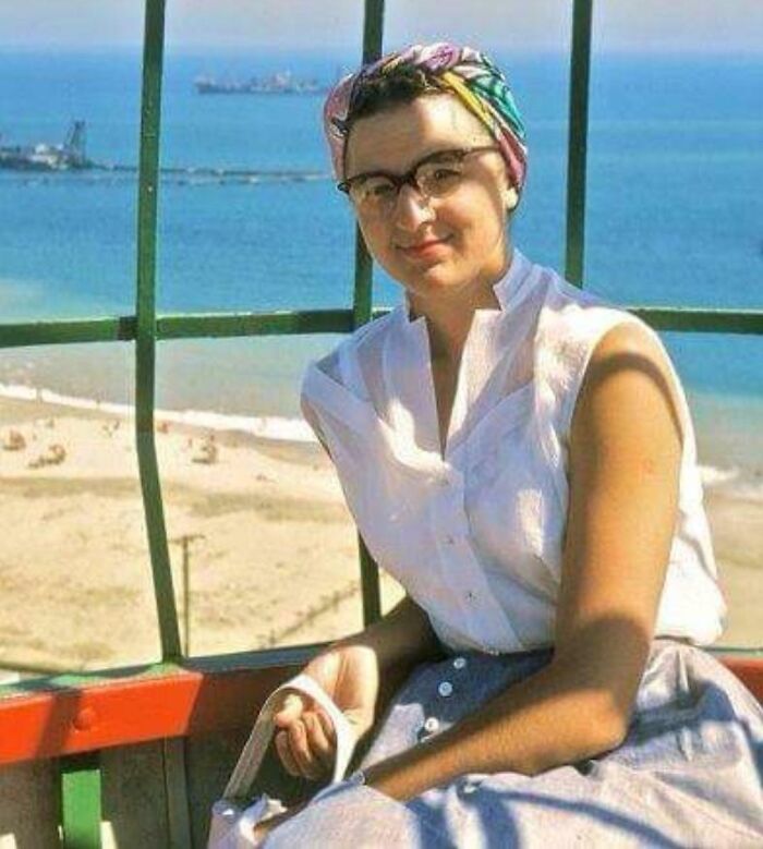 Woman in 60s fashion with a headscarf and glasses, seated by the seaside, wearing a sleeveless blouse and skirt.