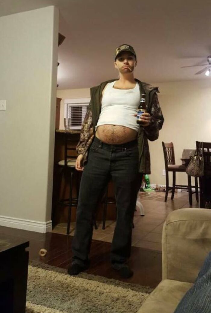 Person humorously dressed with a fake pregnancy belly, holding a beer in a casual home setting.