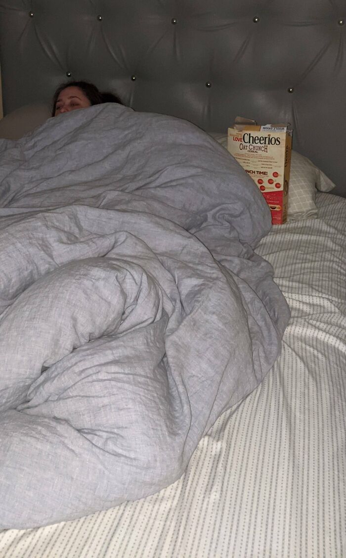 Pregnant woman in bed snuggling under gray blanket with a cereal box beside her, showcasing funny pregnancy situations.
