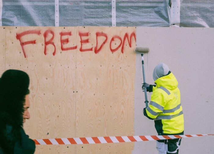 Worker covers graffiti reading "FREEDOM" with paint; scene of creative street photography.