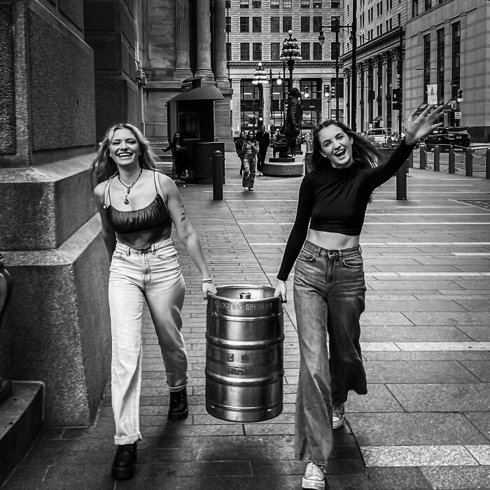 Two women carrying a keg, laughing and walking in an urban setting, showcasing creative street photography.