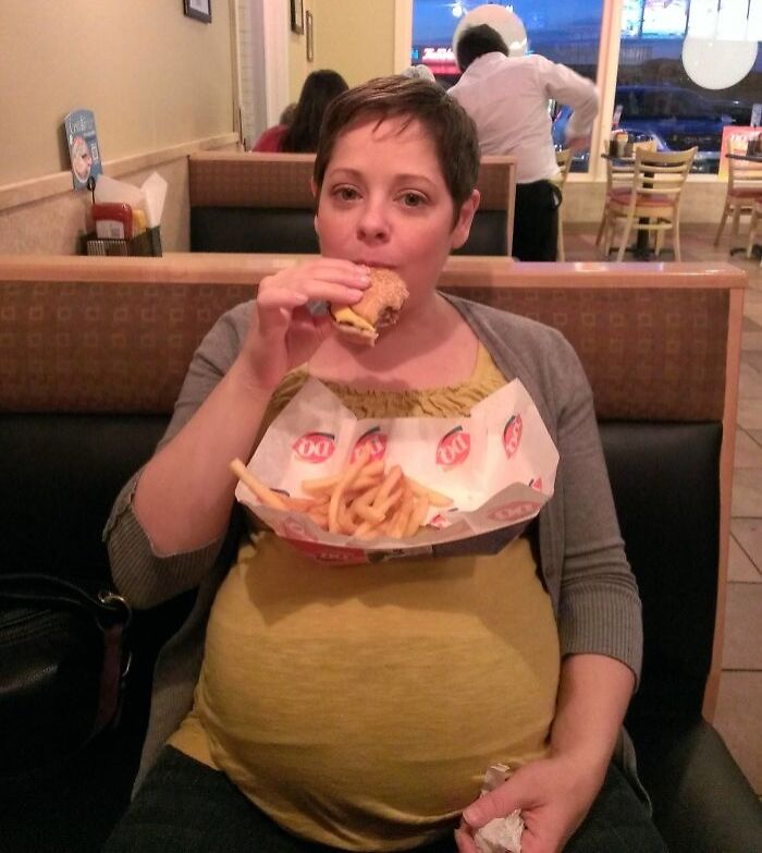 Pregnant woman humorously balancing fast food on her belly at a restaurant.