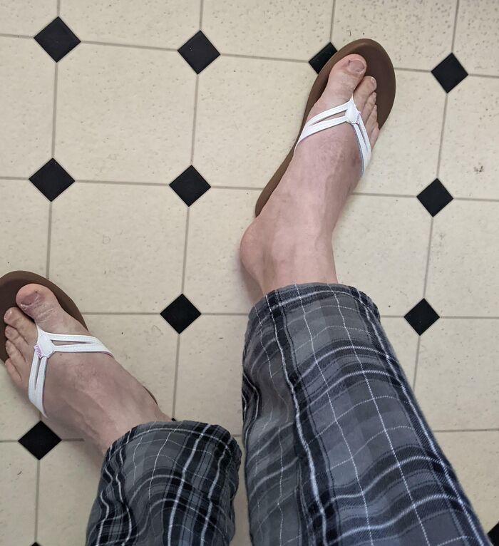 Bare feet wearing flip-flops on a tiled floor, plaid pajama pants visible, capturing a funny pregnancy moment.