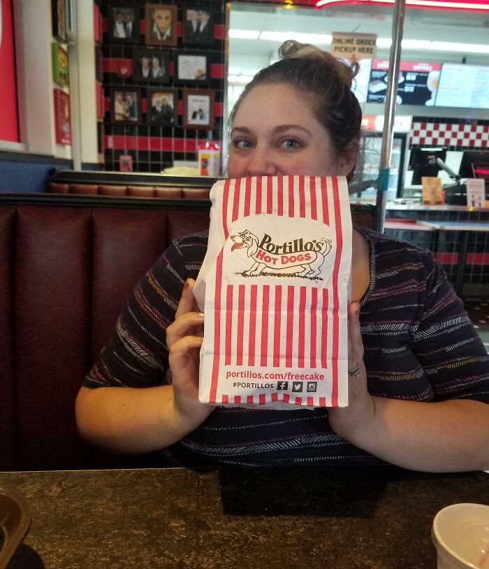 A woman in a restaurant humorously hides her face behind a Portillo's hot dog bag, showcasing funny pregnancy situations.