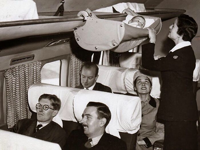 Flight attendant adjusts baby hammock over passengers in vintage airplane cabin, showcasing strange inventions.