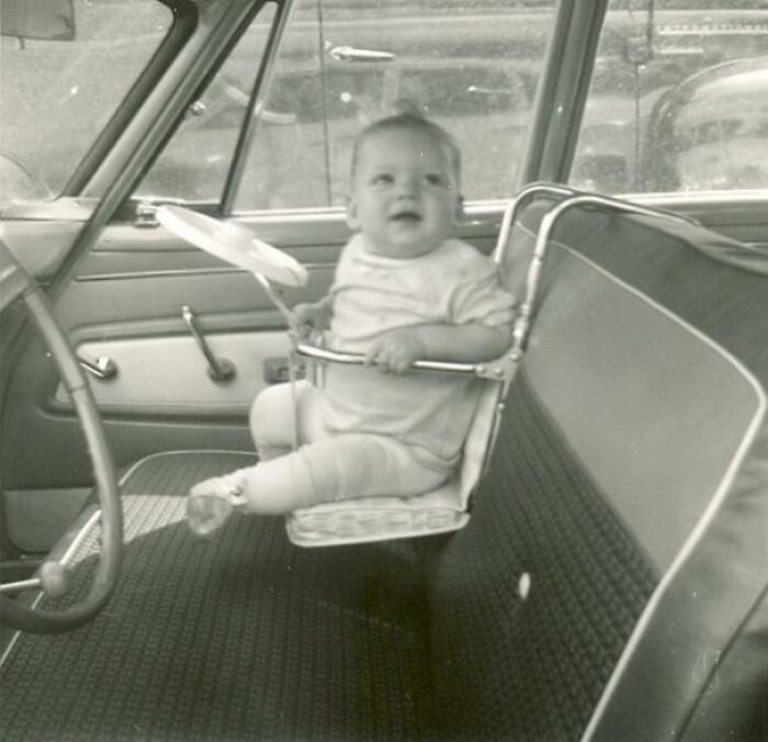 Baby in a strange invention car seat hanging over a bench seat inside a vintage car.