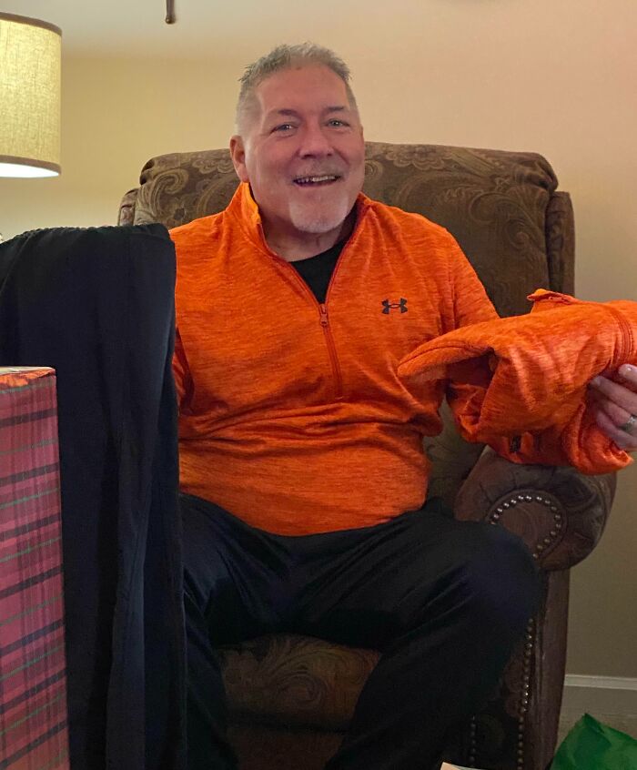 Man in an orange sweater laughing, holding a funny Christmas gift in a cozy living room setting.