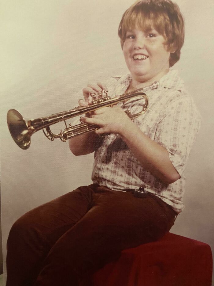 A 1970s high school student with a trumpet, wearing a patterned shirt and brown pants, sitting on a red stool.
