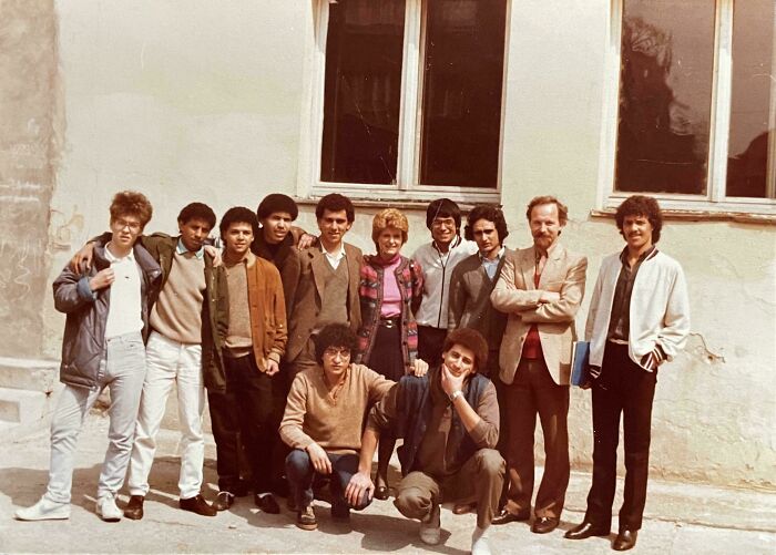 Group of people outside a 1970s high school building, showcasing vintage fashion and hairstyles typical of the era.