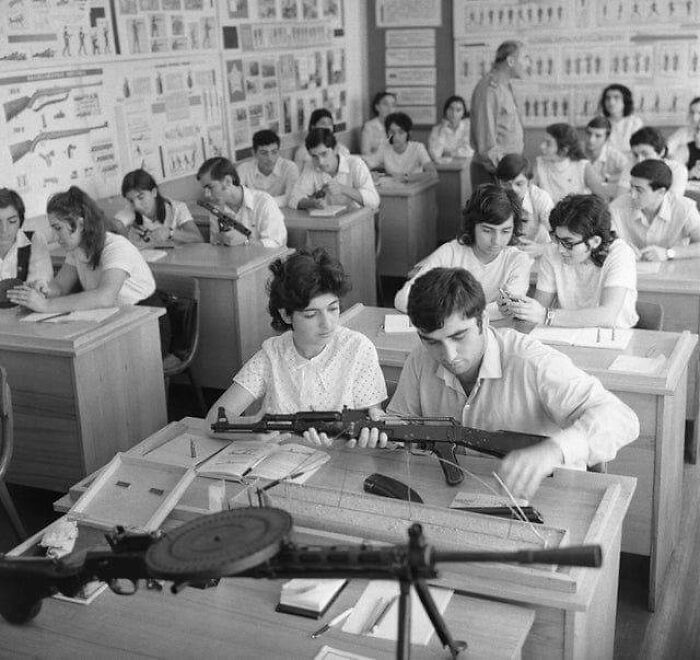 Students in a 1970s high school classroom handling disassembled rifles, with educational posters on the walls.