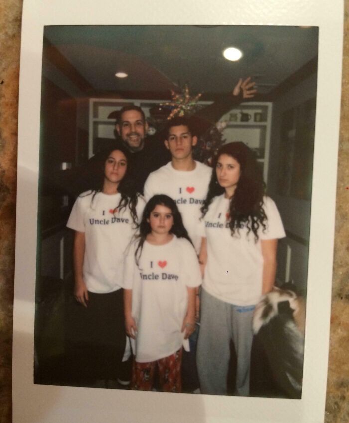 Family wearing matching T-shirts with a humorous message in front of a Christmas tree, showcasing a hilarious trolling gift.