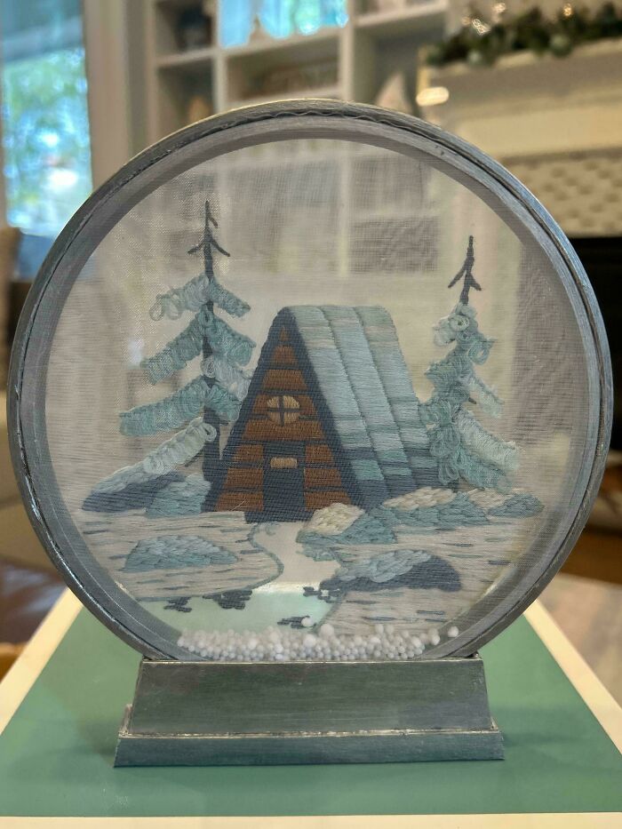Embroidered winter cabin scene with snow-covered trees, detailed stitching inside a round frame on a green base.