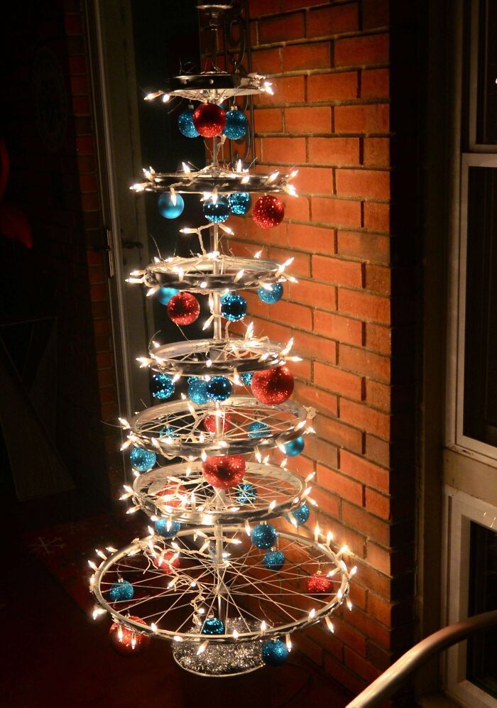 Creative Christmas tree made from bicycle wheels, adorned with red and blue ornaments, against a brick wall.