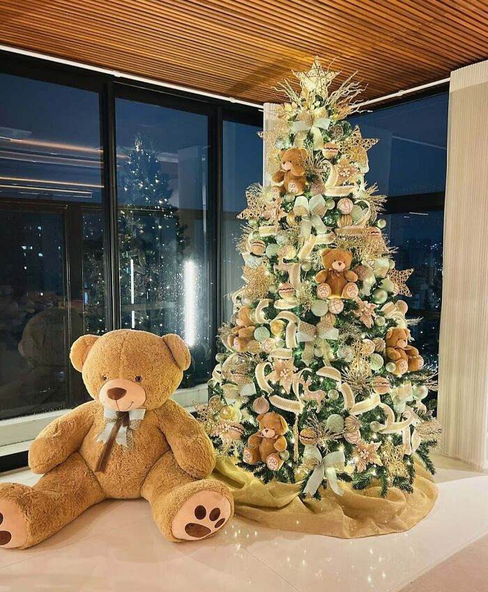 Christmas tree decorated with teddy bears, gold ornaments, and white lights next to a large teddy bear.