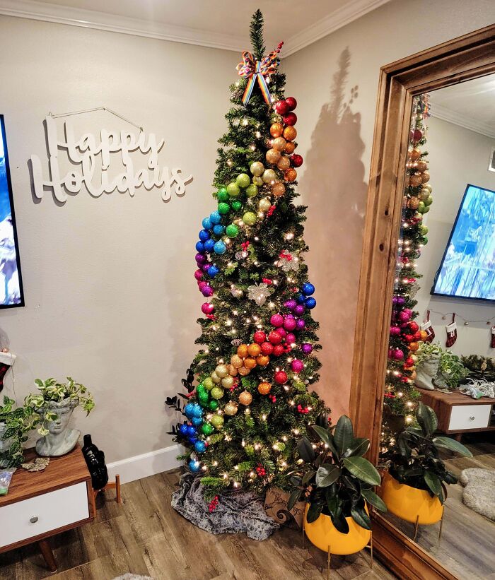 Tall Christmas tree decorated with colorful baubles and lights, standing next to a "Happy Holidays" sign in a cozy living room.