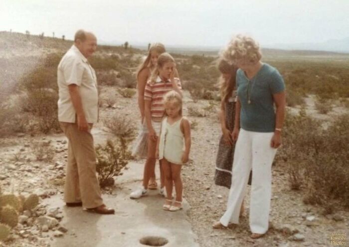 Awkward family photo in a desert, adults and children curiously inspecting a hole in the ground, capturing a vintage moment.