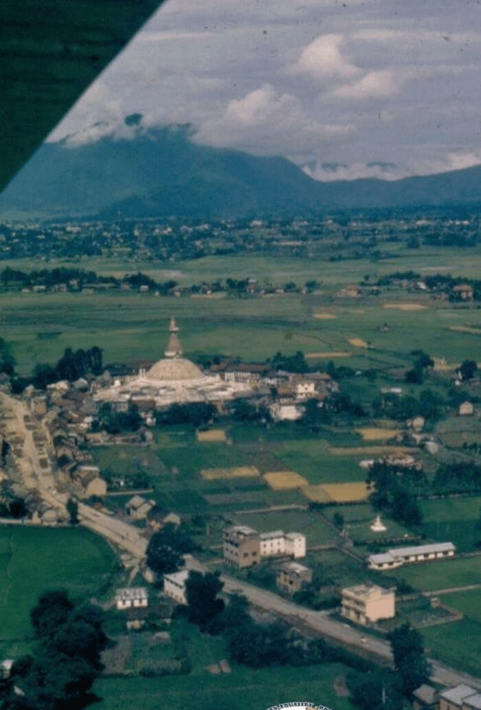 Aerial view of urban landscape with historic stupa, surrounded by fields and buildings against a mountainous backdrop.