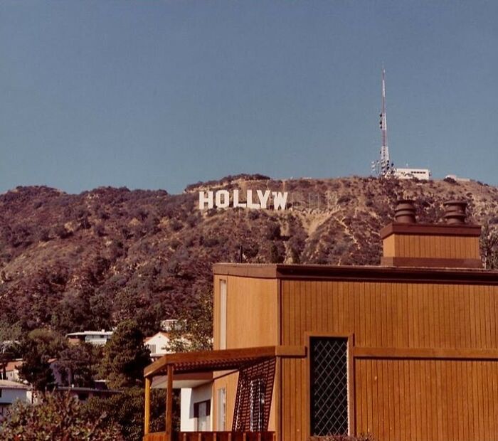 Vintage 1970s West Coast image featuring a partially visible Hollywood sign on a hill with brown wooden buildings below.