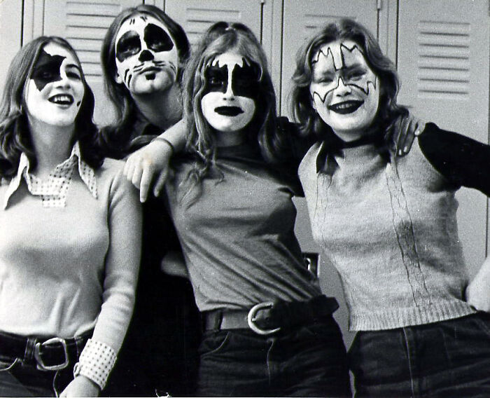 Teens in '70s high school dressed in KISS-style face paint, standing in front of lockers, smiling and posing together.