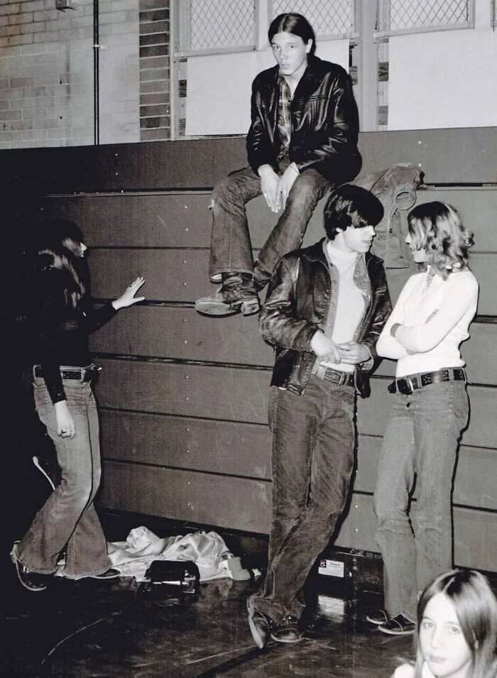 Teens in '70s high school, wearing leather jackets, sitting and standing by gym bleachers, chatting in a relaxed setting.