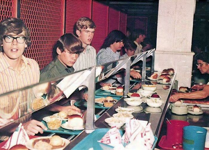1970s high school cafeteria scene with students in line, wearing casual shirts, choosing food trays.