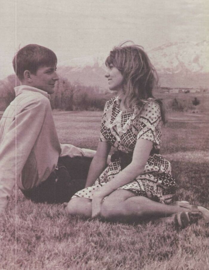 Teen couple in '70s high school fashion, sitting on grass, smiling at each other.