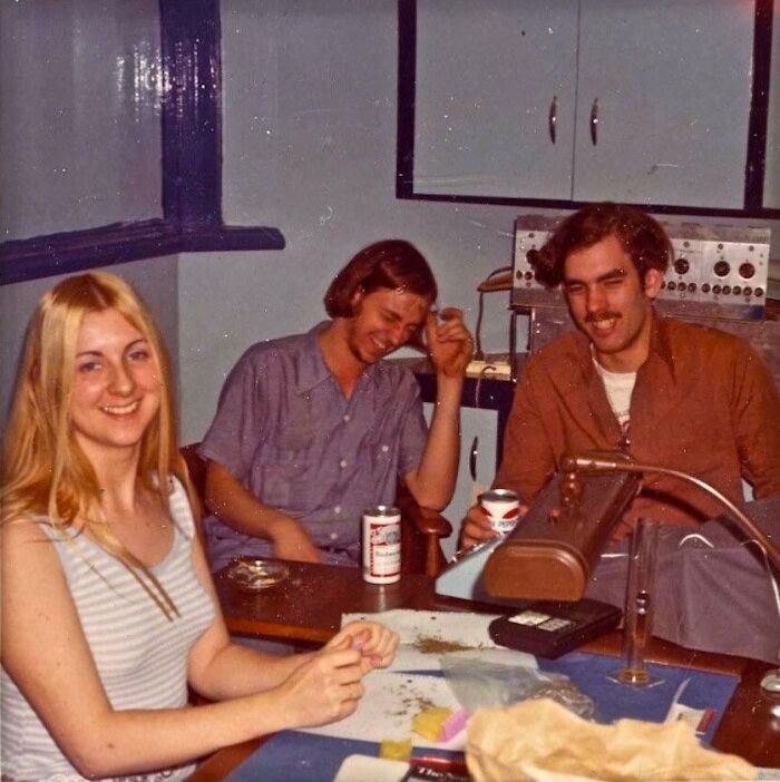 Three friends in a '70s high school classroom, laughing and enjoying drinks, surrounded by vintage equipment.